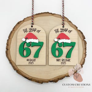 May include: Two wooden Christmas ornaments with the text "THE YEAR OF" and the number "67" in green. Each ornament has a red Santa hat and the names "NICHOLAS 2025" and "MRS. BELFIORE 2025". The ornaments hang from a red and white string.