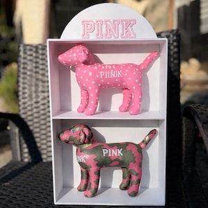 May include: Two plush dog figures in a white display case. The top dog is pink with white polka dots, and the bottom dog is pink and green camouflage. The word "PINK" is printed on both dogs and the display case.