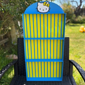 Pode incluir: Uma prateleira de exibição azul e amarela com um design Hello Kitty. A prateleira tem um topo azul com o personagem Hello Kitty e listras verticais amarelas na frente. Está sentada em uma cadeira preta ao ar livre.
