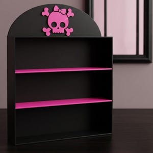 May include: A black wall-mounted shelf unit featuring three tiers with vibrant pink shelves. The top of the unit has a rounded arch shape decorated with a bright pink skull and crossbones icon that is wearing a small bow on its head.