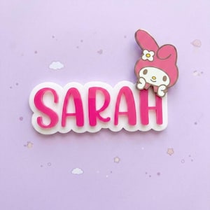 以下が含まれることがあります： 白いスカラップ状の背景に、鮮やかなピンクの太い大文字で「SARAH」と書かれたカスタムネームバッジ。右上隅には、頭に白い花をつけたピンク色の漫画のウサギのキャラクターが取り付けられています。