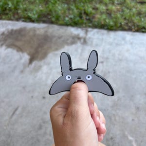 May include: A grey cartoon character with large ears and white eyes. The character is held in a person's hand against a background of concrete and grass. The character has a black outline.