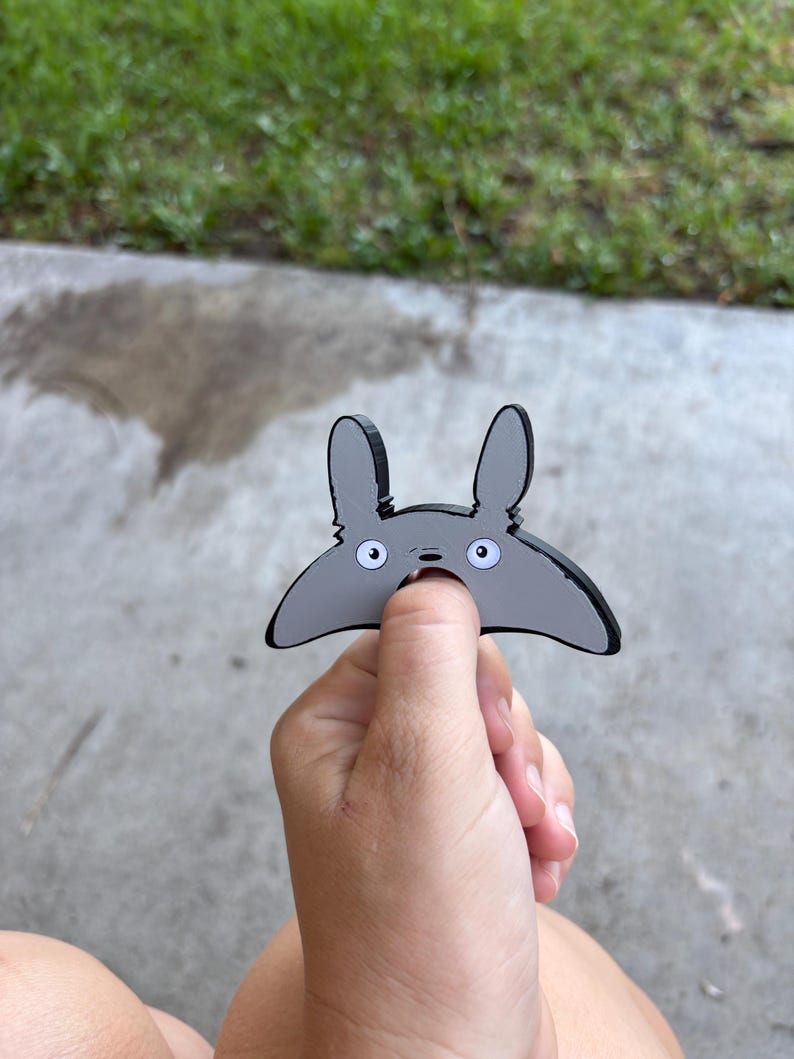 May include: A grey cartoon character with large ears and white eyes. The character is held in a person's hand against a background of concrete and grass. The character has a black outline.