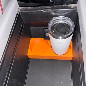 Puede incluir: Un organizador de consola de coche naranja con dos portavasos circulares y una base rectangular. Un vaso aislado blanco con tapa negra se encuentra en el portavasos derecho. El organizador est&aacute; dentro de una consola negra.
