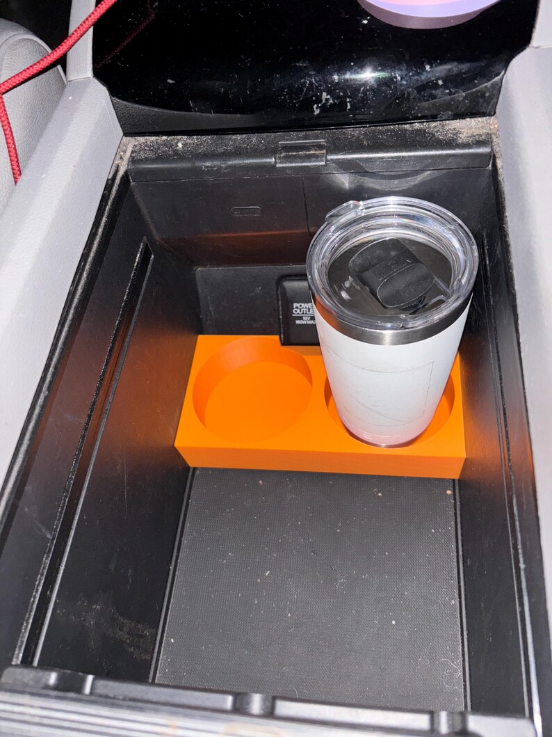 Puede incluir: Un organizador de consola de coche naranja con dos portavasos circulares y una base rectangular. Un vaso aislado blanco con tapa negra se encuentra en el portavasos derecho. El organizador est&aacute; dentro de una consola negra.