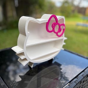 May include: A white Hello Kitty-shaped shelf unit with a pink bow detail. The shelf has three internal levels. The unit is displayed on a black surface, with a blurred green garden in the background.