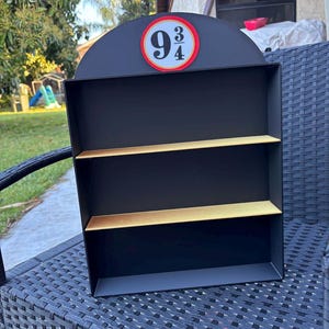 May include: Black display shelf with three levels and a rounded top. The top features a red circle with the numbers 9 and 3/4 in white. The shelves are a light gold color.