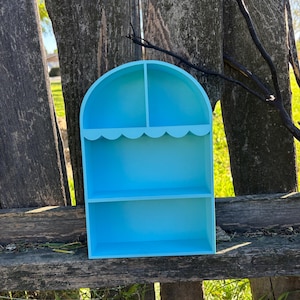 Könnte beinhalten: Hellblaues Holzregal mit gewölbtem Oberteil und gewelltem Rand. Das Regal hat zwei Ebenen und einen geteilten oberen Bereich. Das Regal wird im Freien vor einem verwitterten Holzzaun ausgestellt.