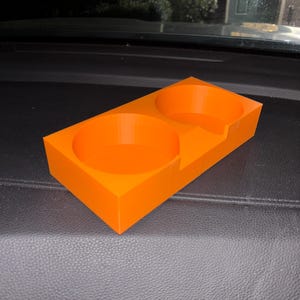 Puede incluir: Portavasos de coche de color naranja brillante con dos aberturas circulares y una base rectangular. El portavasos est&aacute; hecho de un material s&oacute;lido y est&aacute; dise&ntilde;ado para sostener dos bebidas. El dise&ntilde;o es simple y funcional.