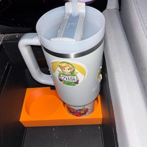 Puede incluir: Un termo de viaje blanco con asa y pajita, adornado con pegatinas de un personaje de dibujos animados y otros dise&ntilde;os. El termo est&aacute; colocado en un portavasos naranja dentro de la consola de un coche.