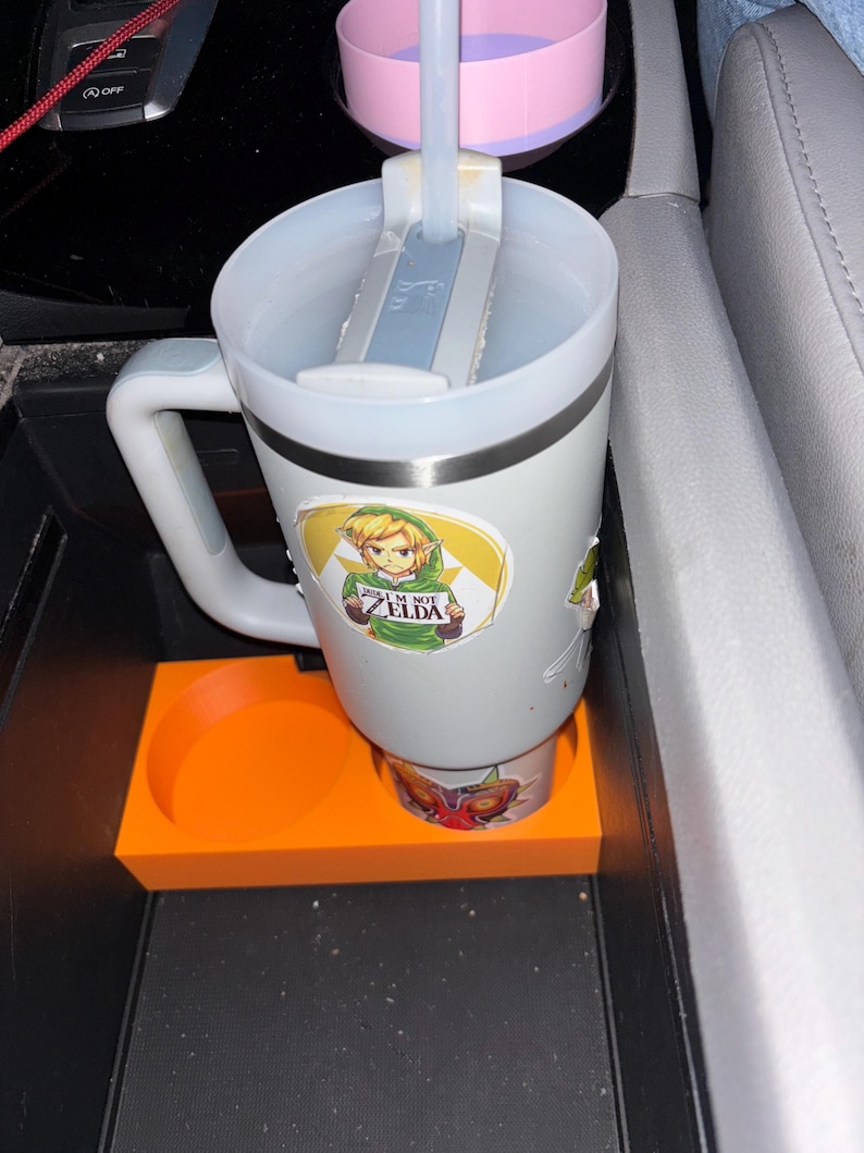 Puede incluir: Un termo de viaje blanco con asa y pajita, adornado con pegatinas de un personaje de dibujos animados y otros dise&ntilde;os. El termo est&aacute; colocado en un portavasos naranja dentro de la consola de un coche.