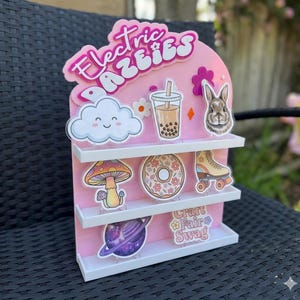 Op de afbeelding: Een roze acryl displaystandaard met drie planken en de tekst Electric Dazzles bovenaan. De standaard is versierd met diverse stickers, waaronder een lachende wolk, een boba-thee, een konijn, een paddenstoel, een donut, een rolschaats, een planeet en de tekst Craft Fair Swag.