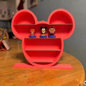 Op de afbeelding: Een felrode Mickey Mouse-vormige plank met drie planken en een voet. De plank bevat drie kleine beeldjes van stripfiguren. De plank is gemaakt van een stevig materiaal en staat op een houten tafel.