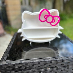 May include: A white Hello Kitty-shaped shelf with a vibrant pink bow. The shelf has two levels for displaying small items. The design is set against a blurred outdoor background, with the shelf resting on a black table.