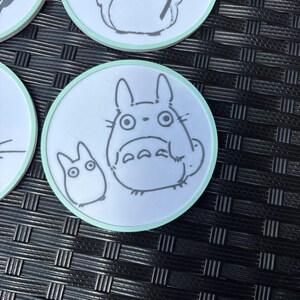 May include: A set of circular white coasters with a light mint green border. Each coaster features a black line art illustration of a large round creature with pointed ears and a smaller companion creature, both depicted in a simple, clean style.