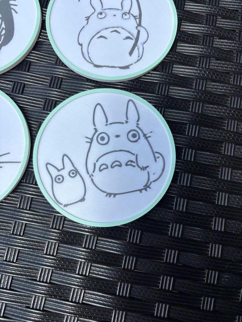 May include: A set of circular white coasters with a light mint green border. Each coaster features a black line art illustration of a large round creature with pointed ears and a smaller companion creature, both depicted in a simple, clean style.