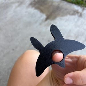 May include: A black metal star-shaped object with a circular hole and two upward protrusions. The object is held in a hand. The background is blurred.