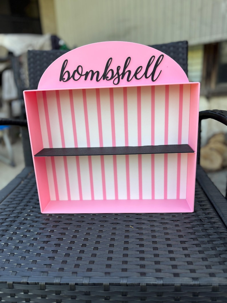Puede incluir: Una estanter&iacute;a rosa con una parte superior semicircular y dos estantes. La palabra "bombshell" est&aacute; escrita en escritura negra en la parte superior. El interior presenta rayas verticales rosas y blancas.