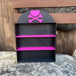 Puede incluir: Una estantería negra con tres estantes, cada uno con un borde rosa. El estante superior tiene un diseño rosa de corazón y huesos cruzados.