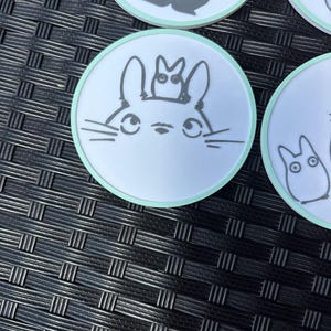 May include: A circular white coaster with a light green border featuring a black line drawing of a large creature with long ears and a smaller creature perched on its head. The design includes whiskers and large eyes, centered on the coaster surface.