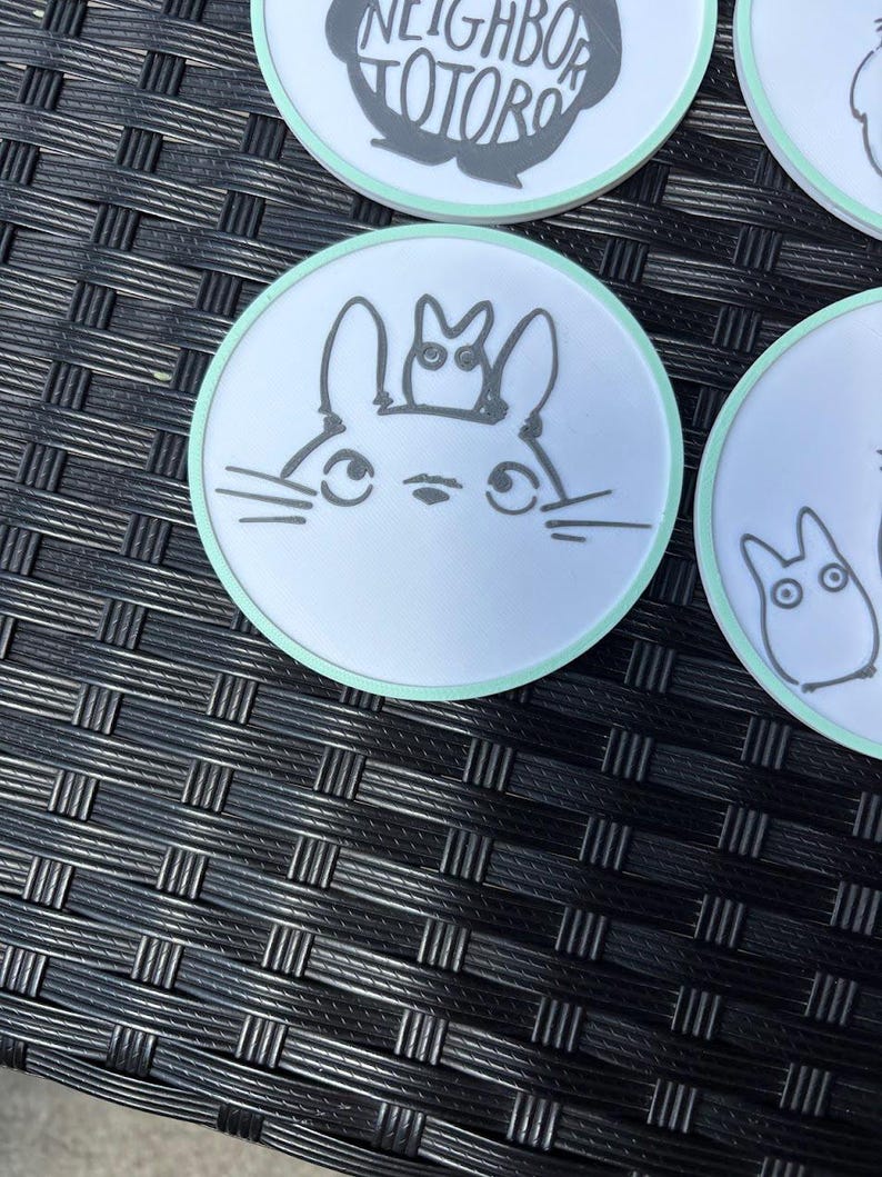 May include: A circular white coaster with a light green border featuring a black line drawing of a large creature with long ears and a smaller creature perched on its head. The design includes whiskers and large eyes, centered on the coaster surface.