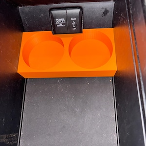 Puede incluir: Portavasos de consola de coche de color naranja brillante con dos aberturas circulares. El portavasos est&aacute; colocado dentro de una consola negra. Encima del portavasos est&aacute;n las palabras "POWER OUTLET" y "AUX".