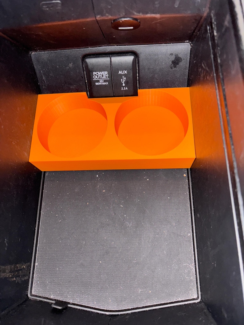 Puede incluir: Portavasos de consola de coche de color naranja brillante con dos aberturas circulares. El portavasos est&aacute; colocado dentro de una consola negra. Encima del portavasos est&aacute;n las palabras "POWER OUTLET" y "AUX".