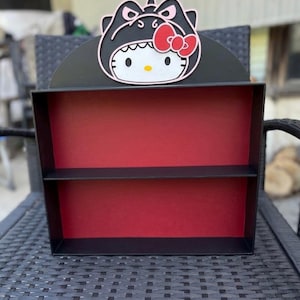 May include: A black wall shelf with a red interior and a single horizontal shelf. The top features a decorative cutout of a white cat character wearing a black dinosaur hood with a red bow, black eyes, and a yellow nose, centered on a black arch.
