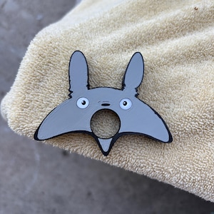 May include: A grey and black Totoro-shaped bottle opener. The bottle opener features a distinctive design with large eyes and pointed ears. The product is made of plastic and is designed to open bottles.