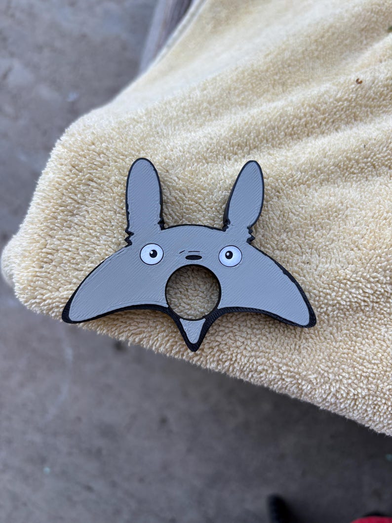 May include: A grey and black Totoro-shaped bottle opener. The bottle opener features a distinctive design with large eyes and pointed ears. The product is made of plastic and is designed to open bottles.