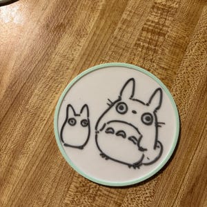 May include: A circular white coaster with a light green border featuring a black line art illustration of a large Totoro character standing next to a smaller Totoro character. The design is printed on a flat, smooth surface with a clean finish.