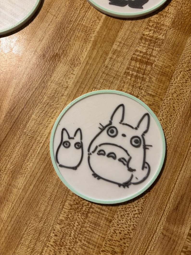 May include: A circular white coaster with a light green border featuring a black line art illustration of a large Totoro character standing next to a smaller Totoro character. The design is printed on a flat, smooth surface with a clean finish.