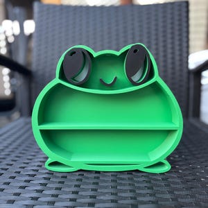 May include: A bright green frog-shaped shelf with two levels. The frog has large black eyes and a smiling mouth. The shelf is made of a solid material and is designed to hold small items. The shelf is sitting on a black woven surface.