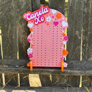 Pode incluir: Um expositor perfurado rosa com o texto Canela Xo em letras laranja e brancas. O painel é decorado com várias formas de corações e flores brancas, laranja e rosa, juntamente com pequenos detalhes de estrelas, apoiado por dois pés laranja.