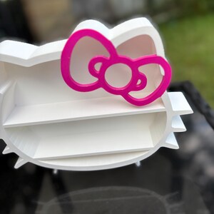 May include: A white, three-tiered shelf shaped like a cartoon cat's head, with a bright pink bow. The shelf has a rounded design with a flat back, suitable for displaying small items. The bow provides a contrasting colour, adding visual interest.