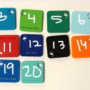 Puede incluir: Una colecci&oacute;n de veinte muestras de color cuadradas de acr&iacute;lico, cada una con un n&uacute;mero escrito en blanco. Las muestras son de varios colores, incluyendo marr&oacute;n, blanco, azul, verde y rosa. Cada muestra tiene un peque&ntilde;o agujero en la esquina superior.