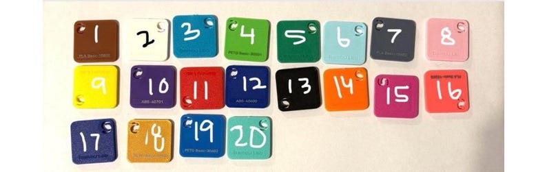 Puede incluir: Una colecci&oacute;n de veinte muestras de color cuadradas de acr&iacute;lico, cada una con un n&uacute;mero escrito en blanco. Las muestras son de varios colores, incluyendo marr&oacute;n, blanco, azul, verde y rosa. Cada muestra tiene un peque&ntilde;o agujero en la esquina superior.