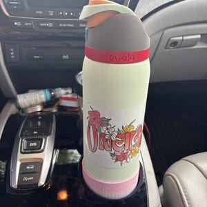 Puede incluir: Una botella de agua Owala verde claro con una banda roja y un diseño floral. La botella está en un portavasos de coche. La botella tiene la palabra "Owala" impresa.