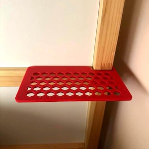 Puede incluir: Una estantería roja brillante con un patrón de panal, fijada a un marco de madera clara. La estantería es de plástico o material similar. El marco de madera forma parte de una cama o mueble similar, sirviendo como pequeño espacio de almacenamiento.
