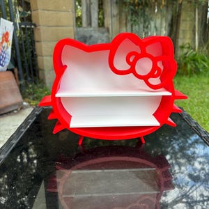 May include: A red, two-tiered shelf shaped like a cartoon cat's head with a bow. The shelves are white, and the design is a playful storage solution. The shelf is on a black table. The overall height is approximately 25 cm.