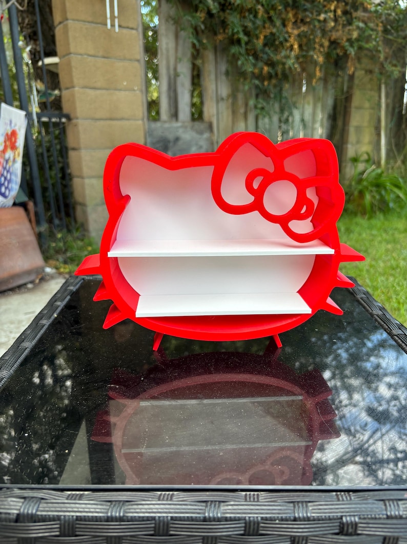 May include: A red, two-tiered shelf shaped like a cartoon cat's head with a bow. The shelves are white, and the design is a playful storage solution. The shelf is on a black table. The overall height is approximately 25 cm.