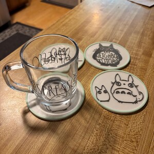 May include: A clear glass espresso mug with a handle and the word ESPRESSO printed in black, resting on a round white coaster featuring a black line drawing of a character. Two additional round coasters with black character illustrations are nearby.