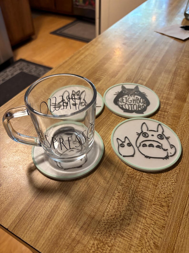 May include: A clear glass espresso mug with a handle and the word ESPRESSO printed in black, resting on a round white coaster featuring a black line drawing of a character. Two additional round coasters with black character illustrations are nearby.
