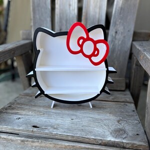 May include: A white Hello Kitty-shaped shelf with a black outline and a red bow. The shelf has two levels and is supported by small white legs. The shelf is placed on a wooden chair.