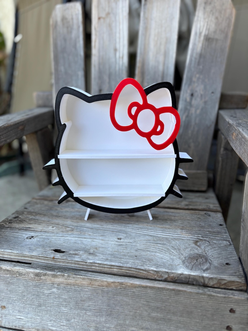 May include: A white Hello Kitty-shaped shelf with a black outline and a red bow. The shelf has two levels and is supported by small white legs. The shelf is placed on a wooden chair.