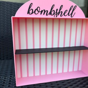 Puede incluir: Una estanter&iacute;a rosa con la palabra "bombshell" en escritura negra. El interior presenta rayas verticales rosas y blancas, con una estanter&iacute;a negra. La estanter&iacute;a est&aacute; dise&ntilde;ada para guardar objetos peque&ntilde;os.