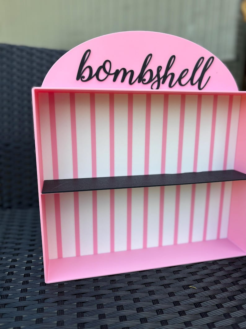 Puede incluir: Una estanter&iacute;a rosa con la palabra "bombshell" en escritura negra. El interior presenta rayas verticales rosas y blancas, con una estanter&iacute;a negra. La estanter&iacute;a est&aacute; dise&ntilde;ada para guardar objetos peque&ntilde;os.