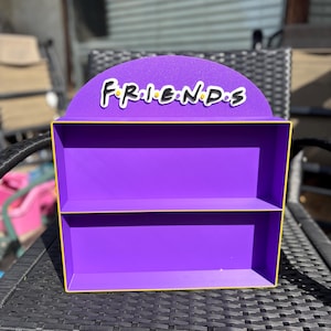 Op de afbeelding: Een paars rek met twee niveaus en een gele rand. De bovenkant van het rek heeft een halfrond ontwerp met het woord "FRIENDS" in witte letters. Het rek staat op een zwarte rotan tafel.