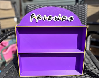 May include: A purple shelf with two levels and a yellow trim. The top of the shelf has a semi-circular design with the word "FRIENDS" in white letters. The shelf is sitting on a black wicker table.