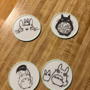 May include: Four circular white coasters with a light mint green border. Each coaster features a black line art illustration of characters from My Neighbor Totoro. One coaster includes the text My Neighbor Totoro in a bold black font.